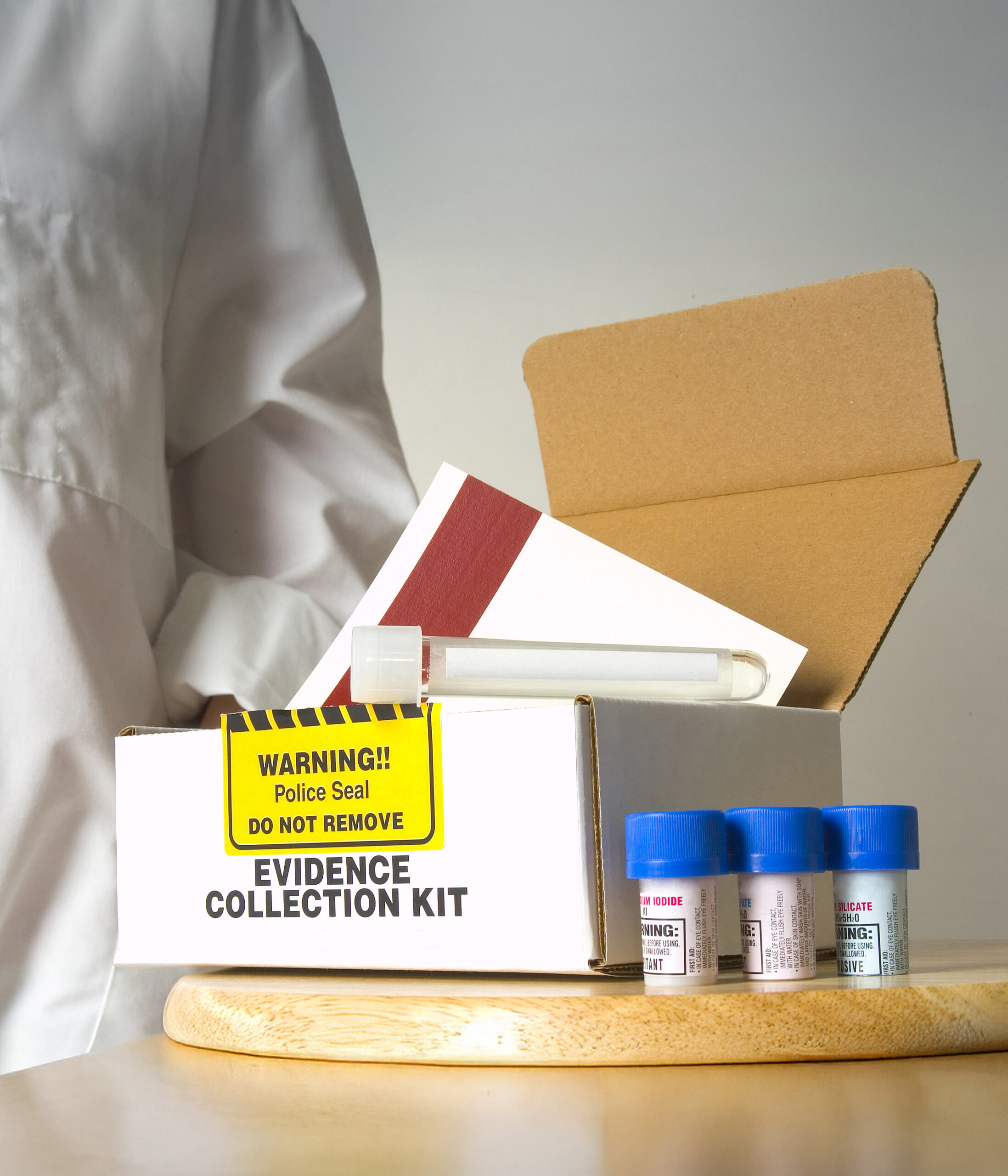 Photo of lab technician standing next to a Sexual Assault Kit. Text on box labels reads "Warning!! Police Seal. Do Not Remove" and "Evidence Collection Kit"