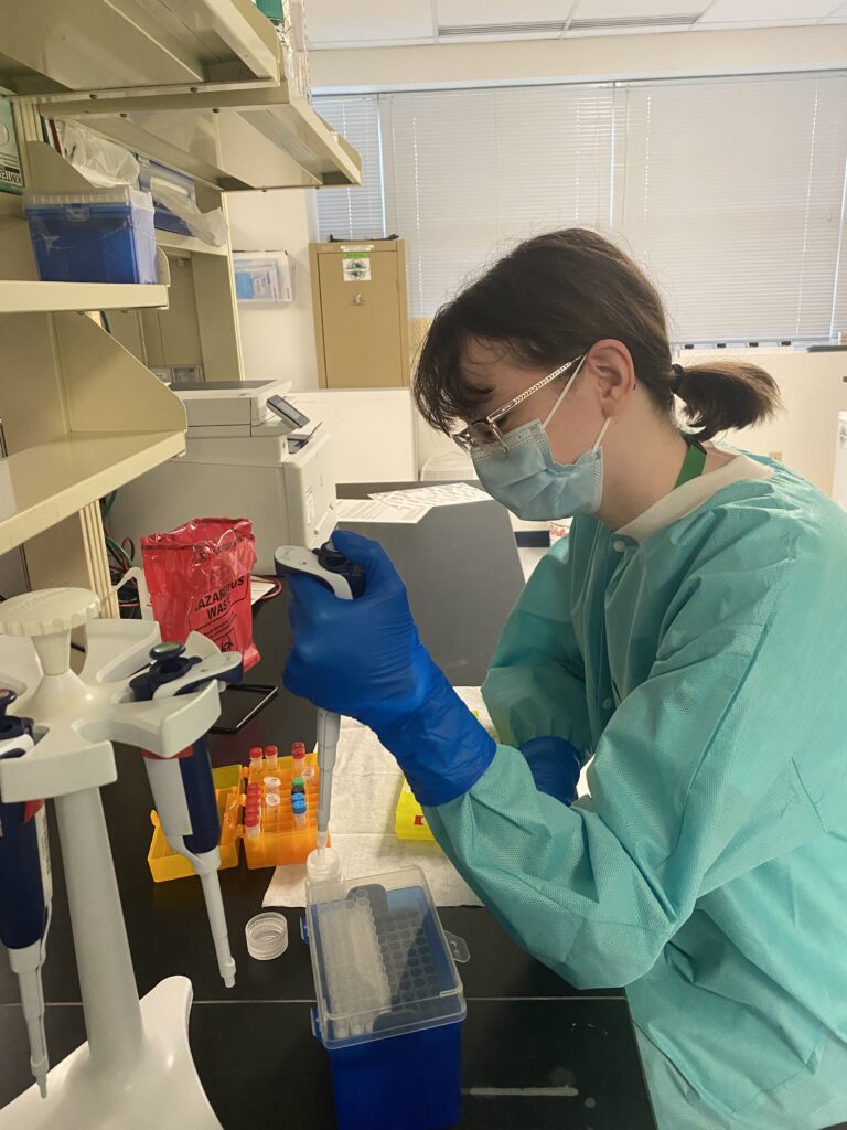 A woman in a lab coat, gloves, and a mask pipettes a solution into a flask.