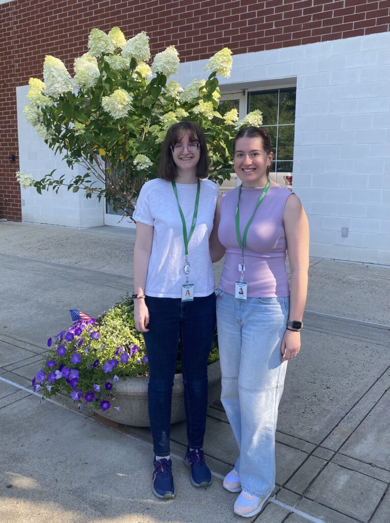 Two young women pose for a photo in front of a flowering tree and brick wall.